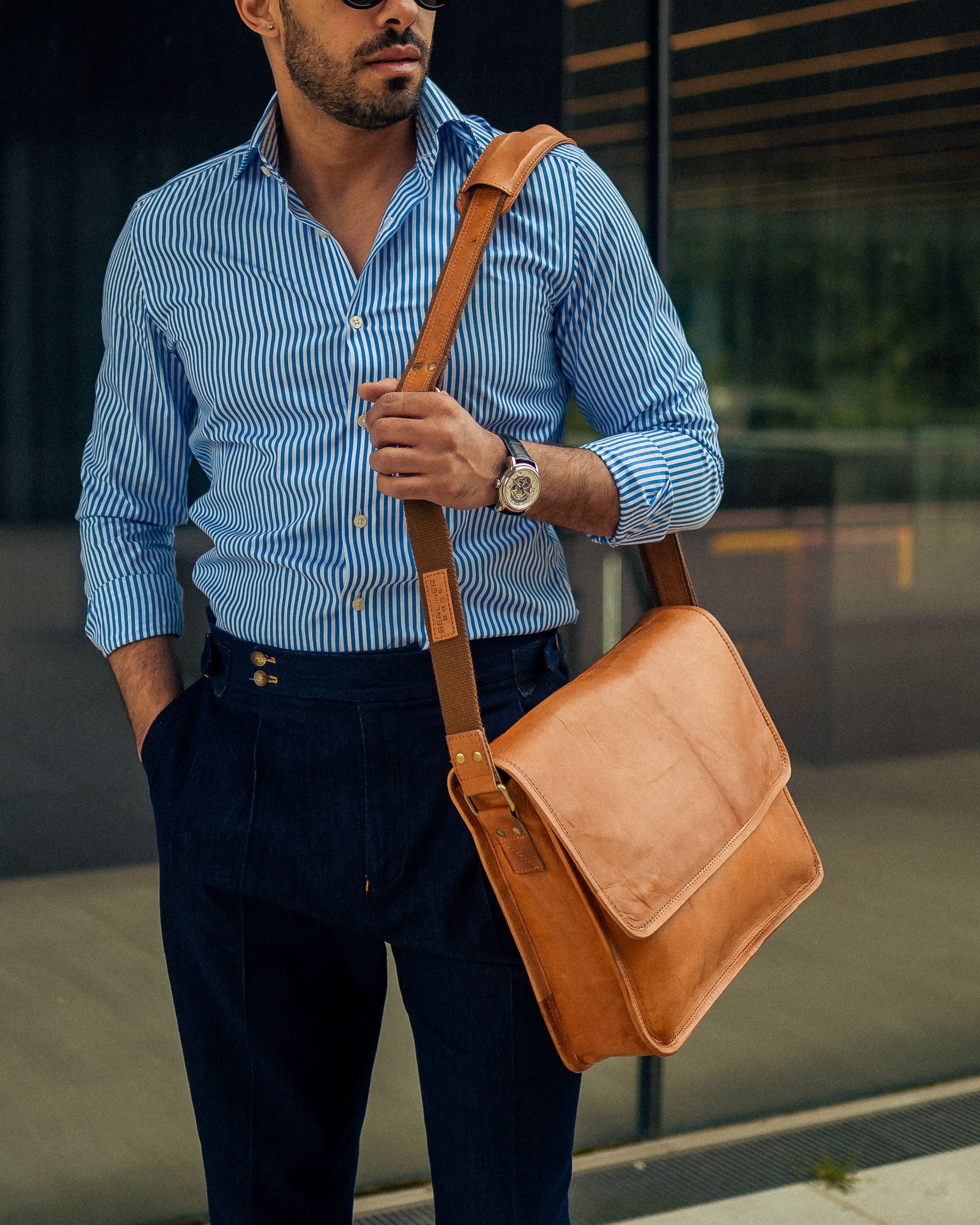Three-quarter view of a man wearing a blue striped dress shirt and dark trousers, holding the shoulder strap of a minimalist vintage leather messenger bag slung across his chest against a dark glass window background