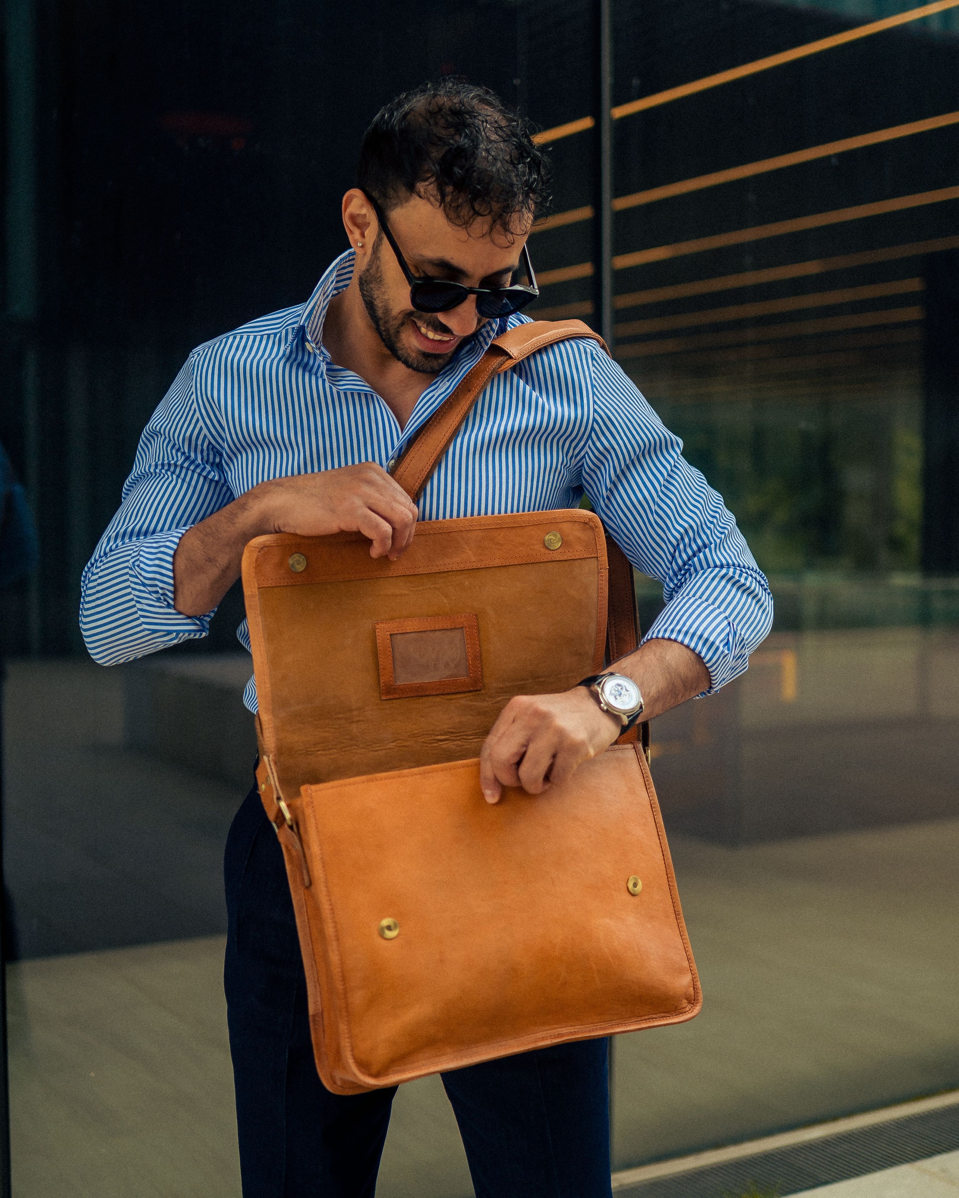 Three-quarter view of a smiling man wearing sunglasses, a blue striped dress shirt, and dark trousers, looking down while opening the main flap of his minimalist vintage leather messenger bag to access the front compartment
