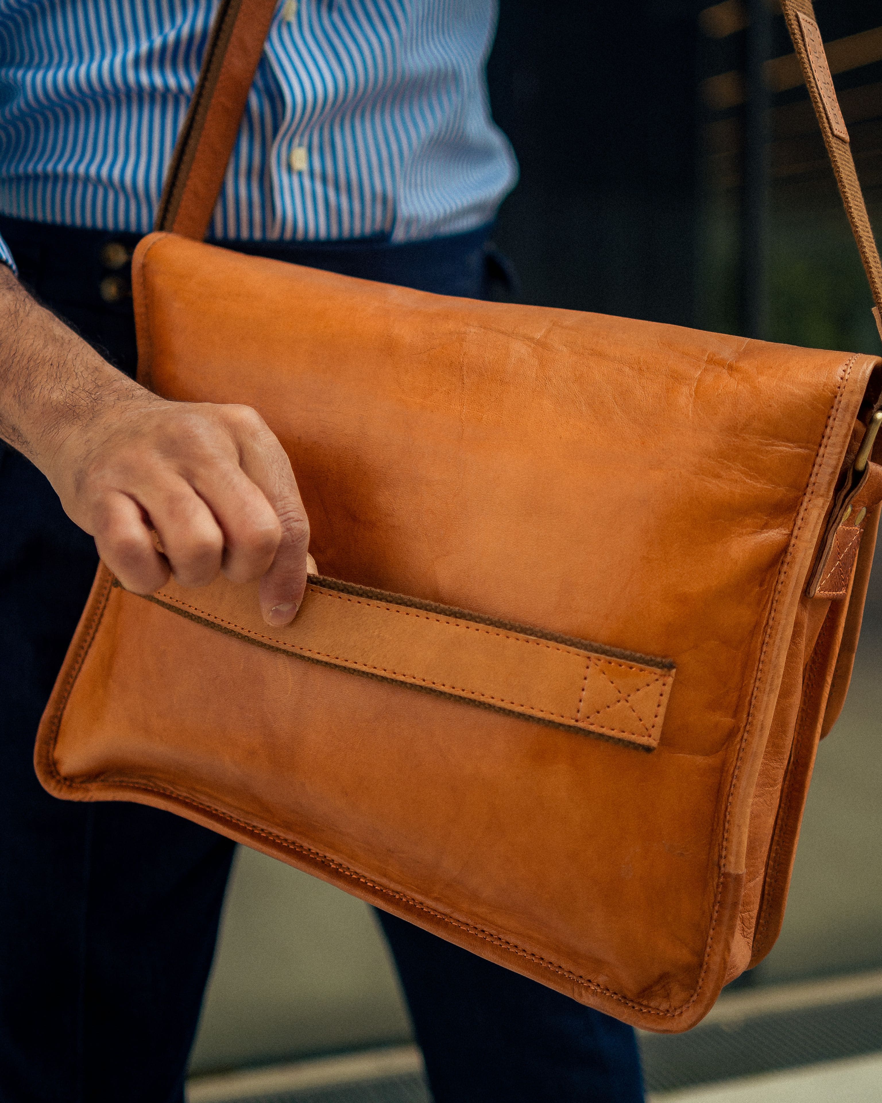 Cropped view of a man wearing a blue striped dress shirt and dark trousers, showing his hand gripping the horizontal luggage trolley strap on the rear side of a minimalist vintage leather messenger bag
