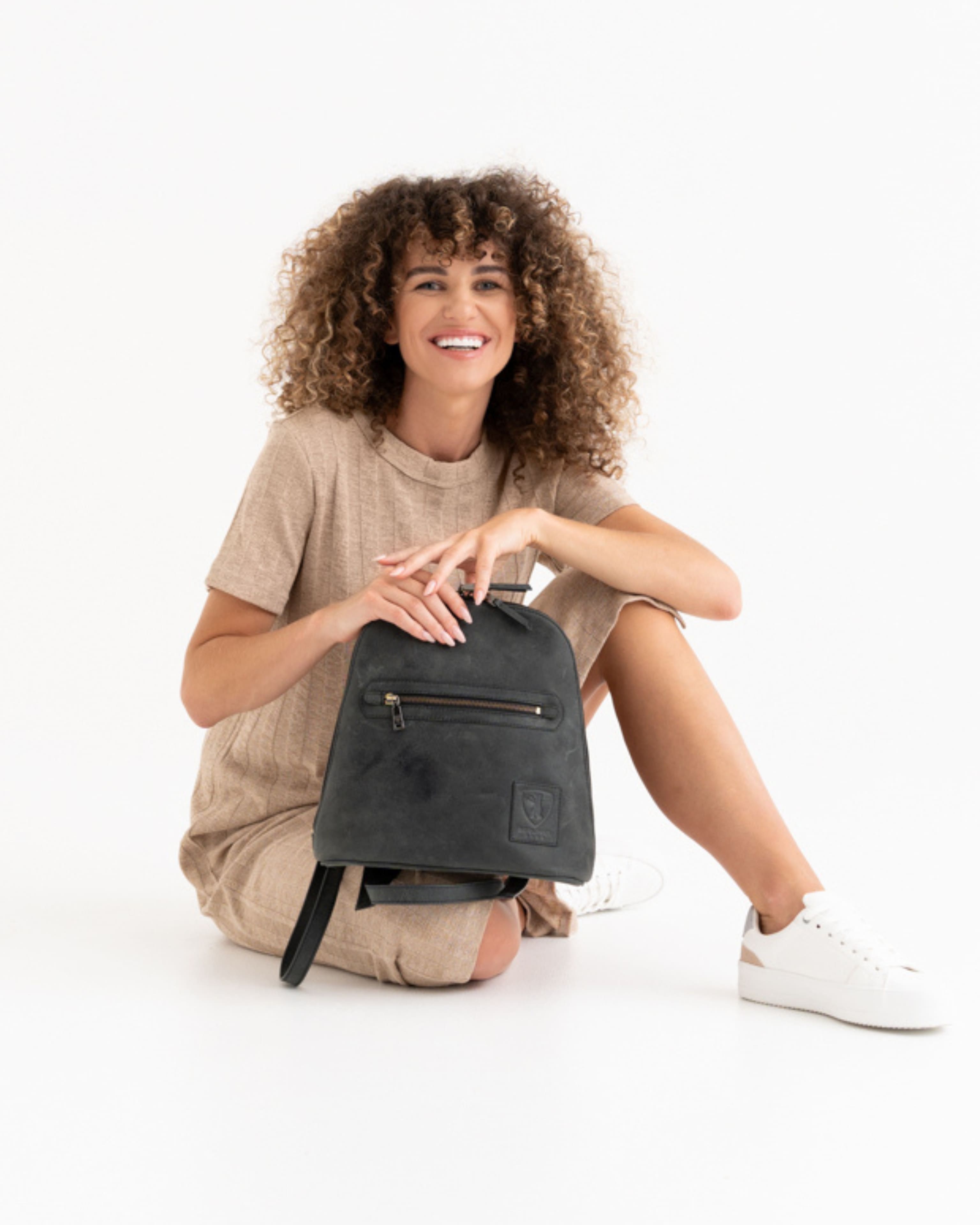 Smiling woman sitting on floor holding dark grey leather backpack