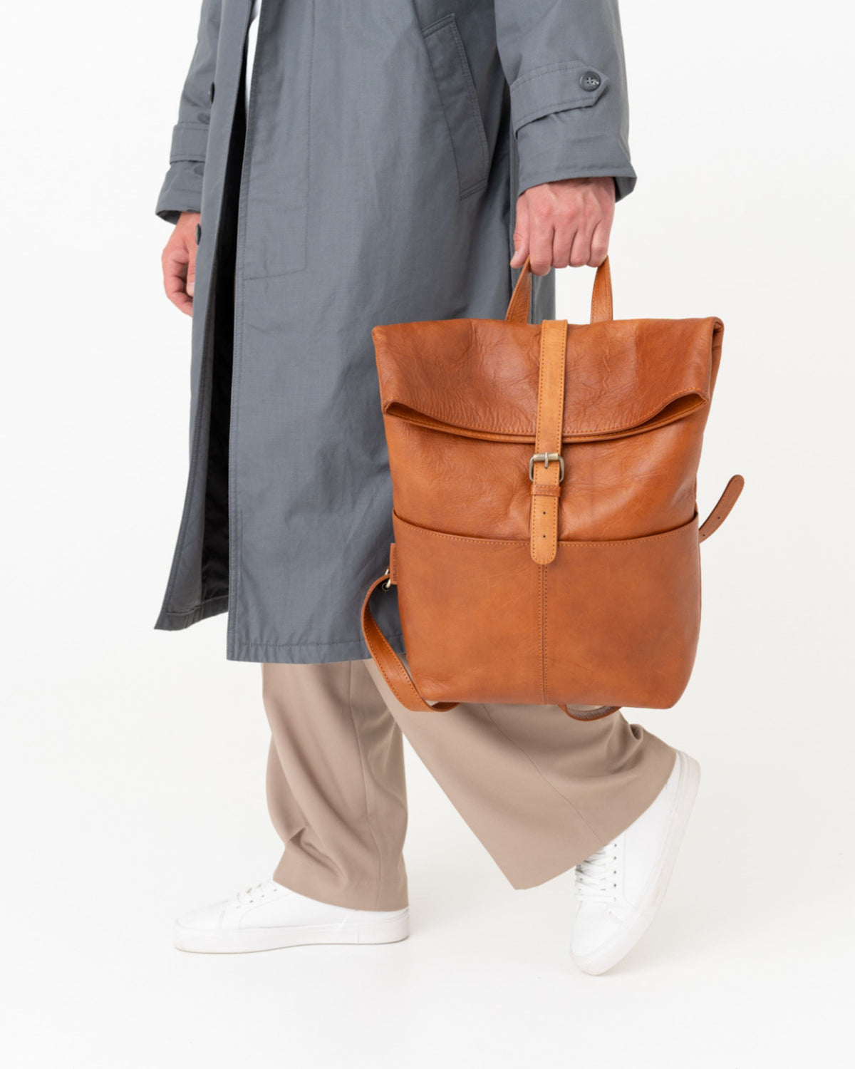 Person in trench coat walking and holding a vintage leather roll-top backpack.