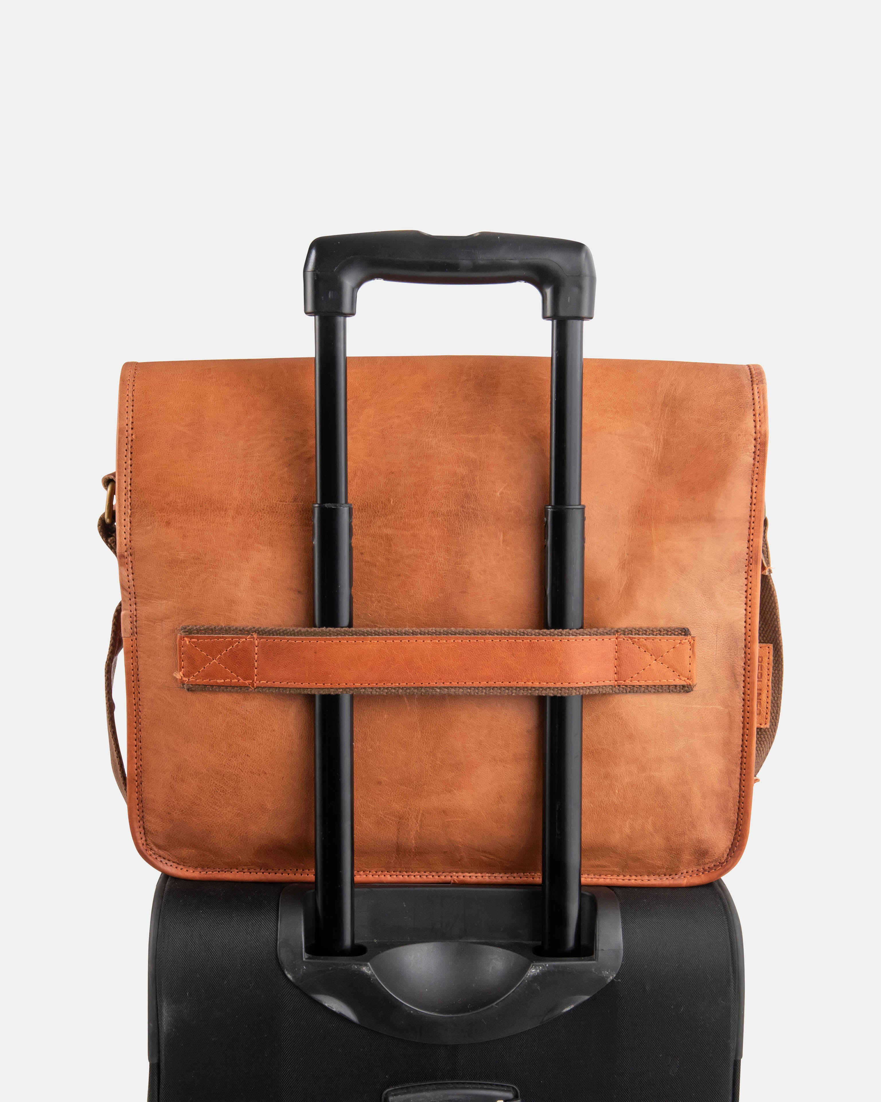 Rear view of a minimalist vintage leather messenger bag from image_9a1329.png, demonstrating the function of its luggage trolley strap by being secured onto the extended handle of a black rolling suitcase, against a plain studio background.