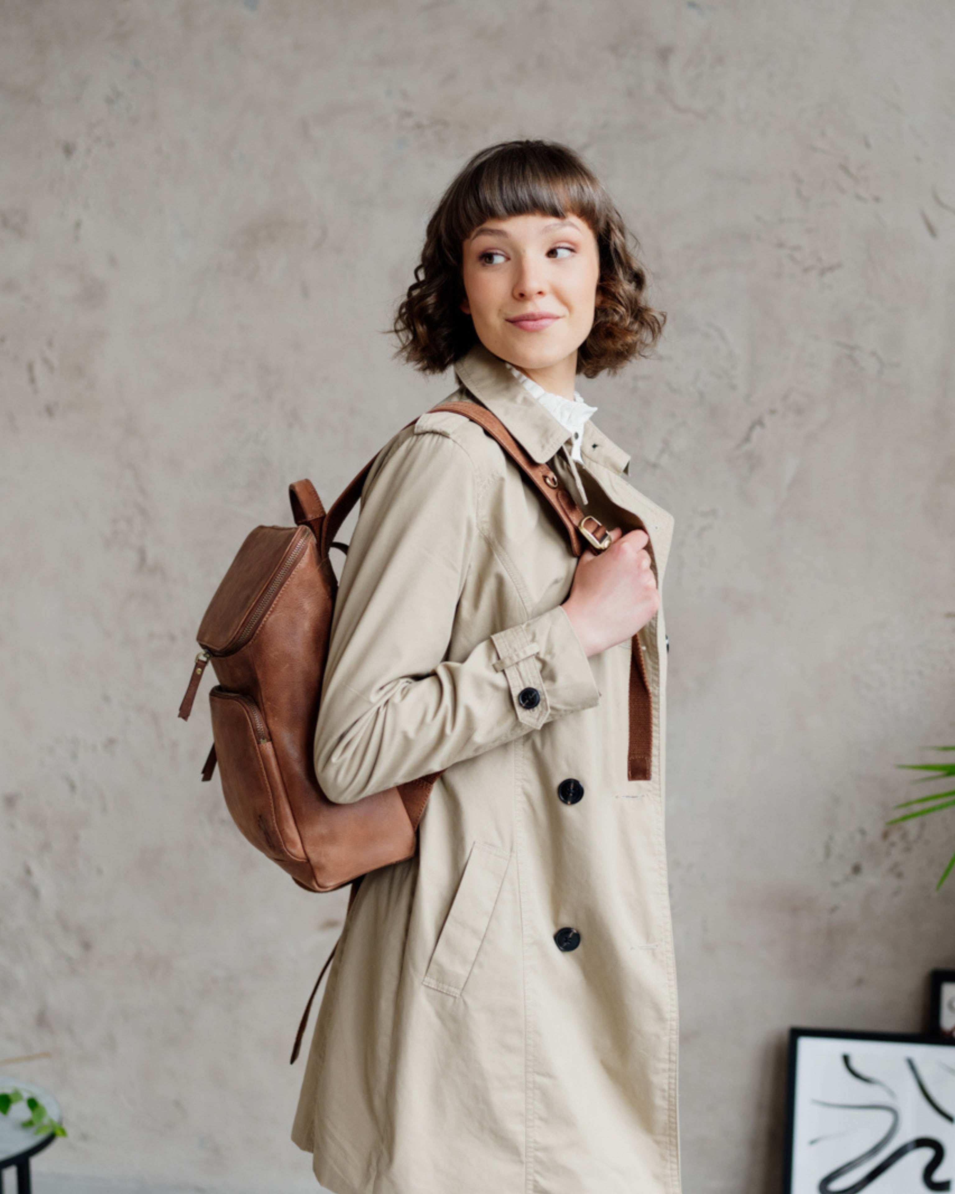 Side lifestyle view of woman in trench coat wearing camel leather backpack.