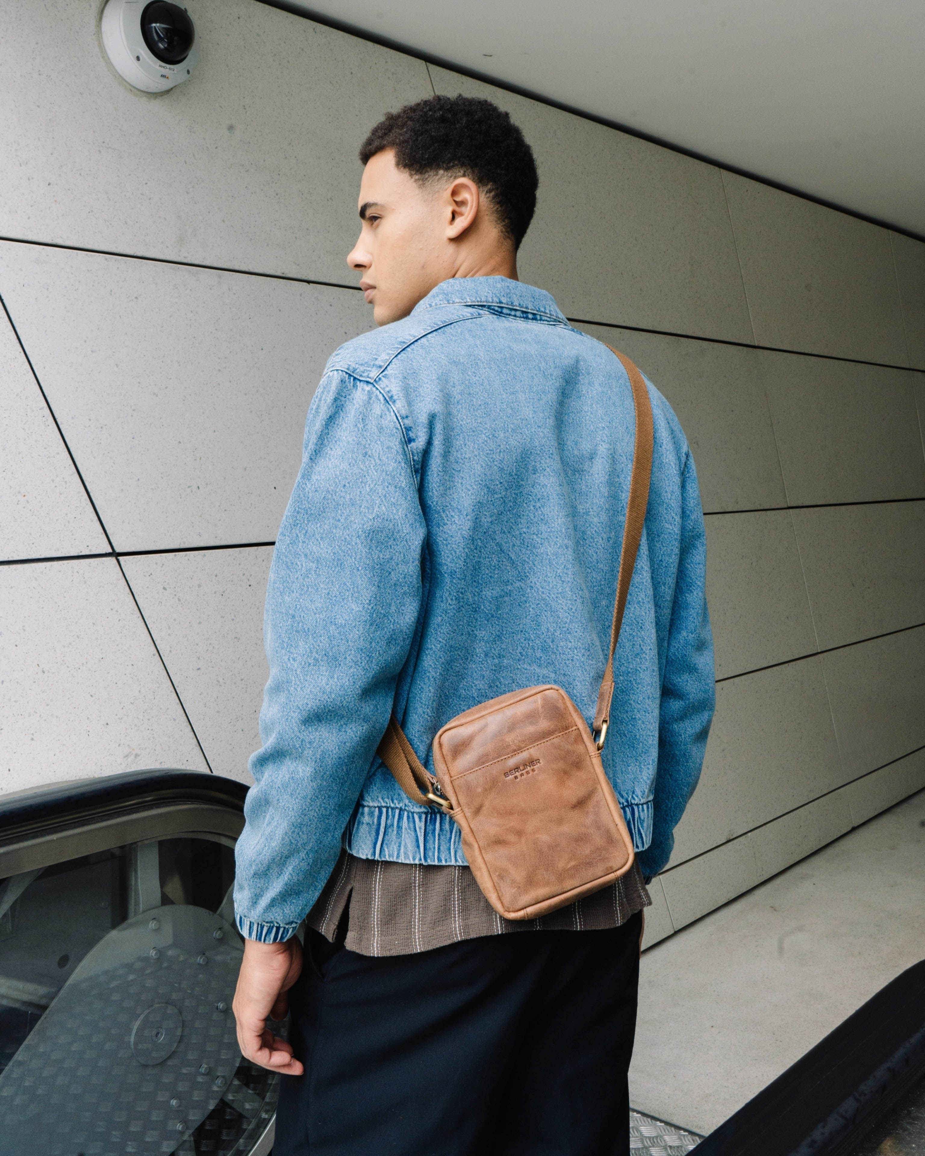 Angled back view of man wearing a denim jacket and a camel leather messenger crossbody bag with a strap