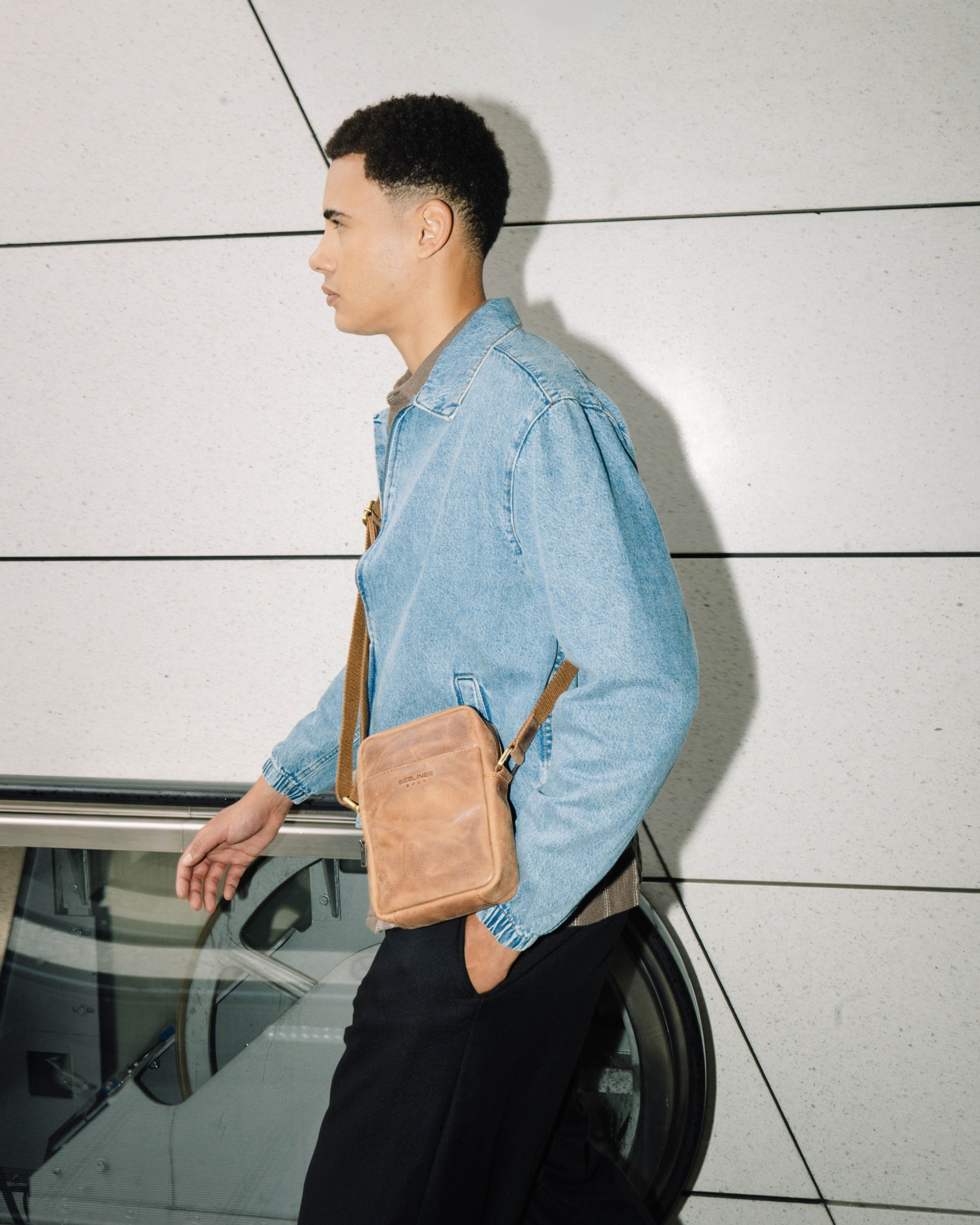 Side profile view of man walking, wearing a denim jacket and a camel leather messenger crossbody bag with a strap