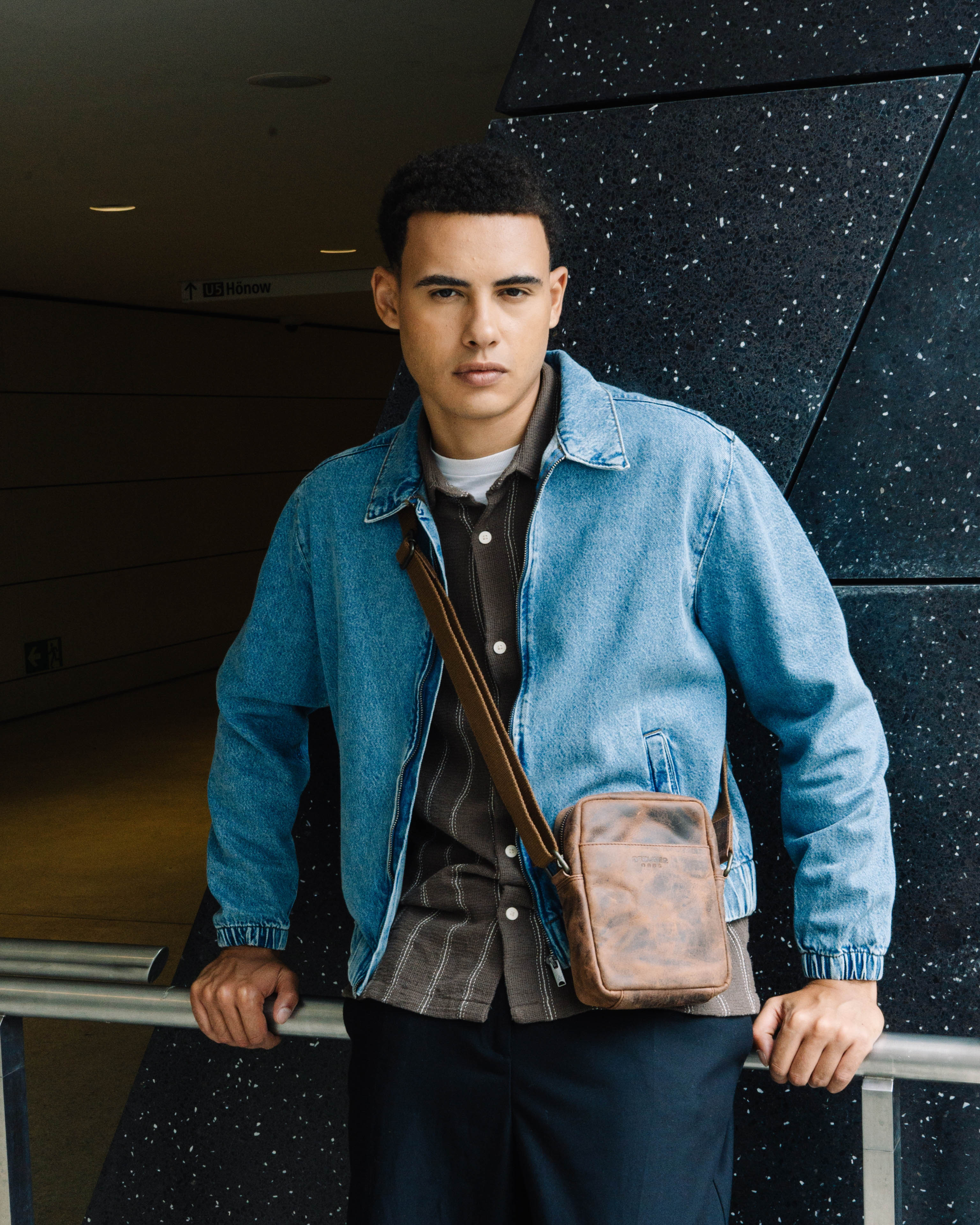 Front view of man wearing a denim jacket and a earth leather messenger crossbody bag with a strap