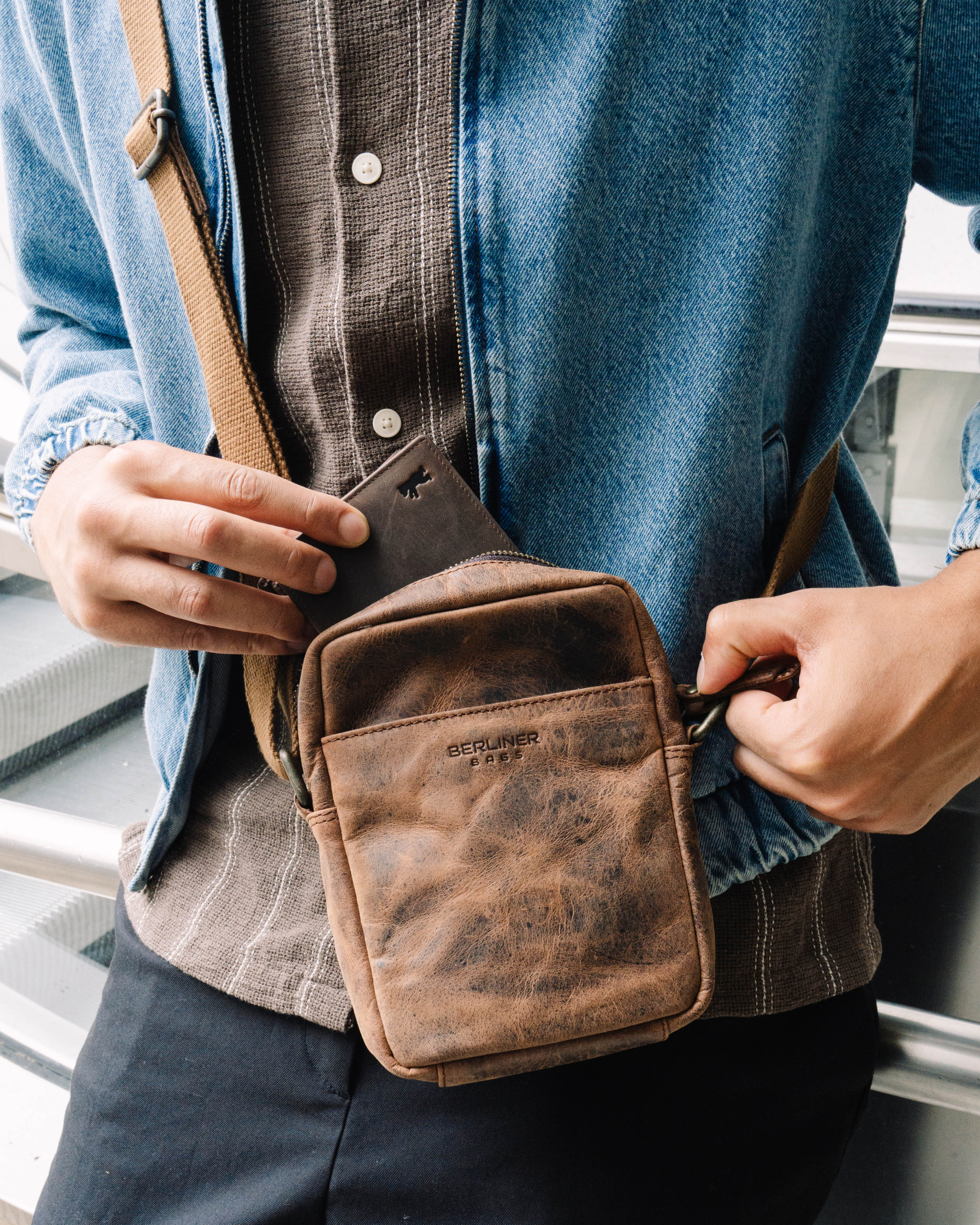 Close up view of man wearing a denim jacket and sliding a wallet into a earth leather messenger crossbody bag