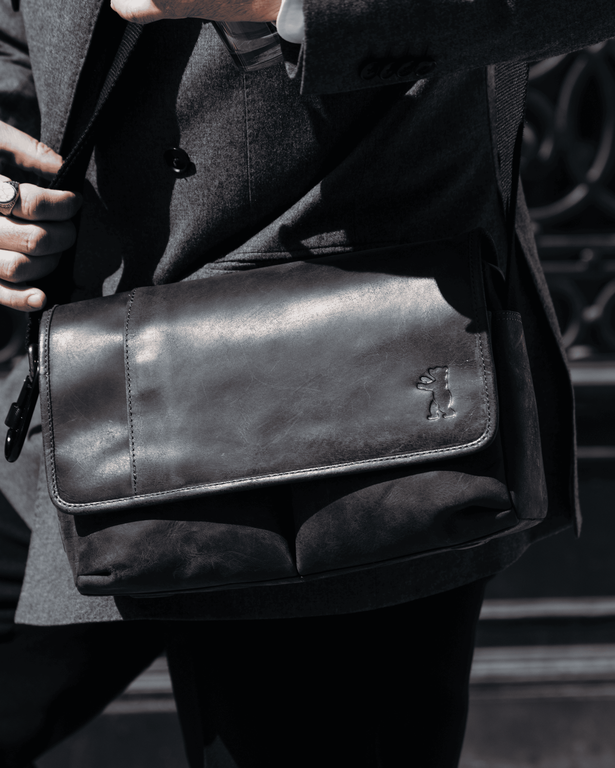 Close-up view of a dark grey leather messenger bag being worn by a person in a grey suit