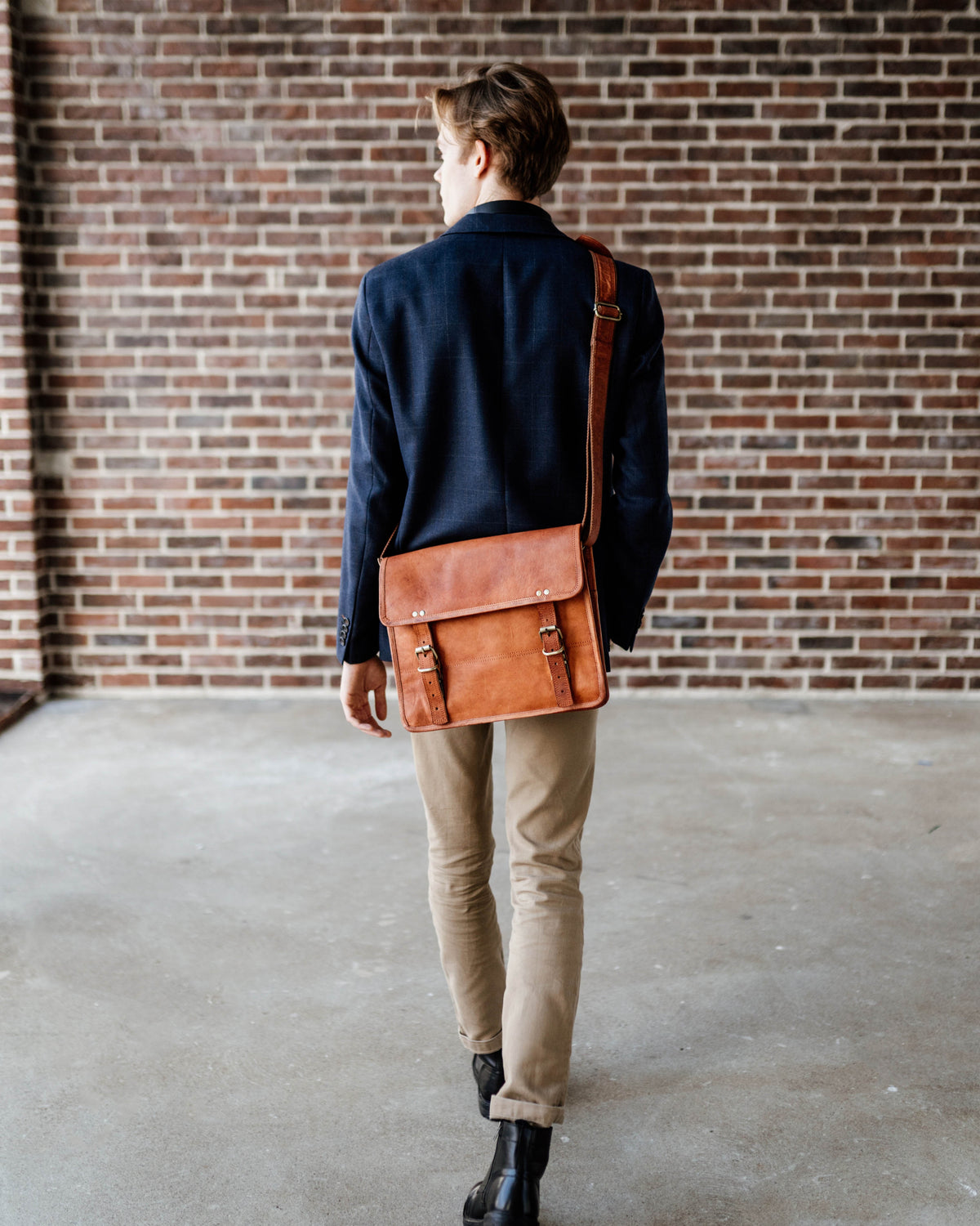 Full-length back view of a man walking away while wearing a navy blazer and khaki pants, carrying a vintage leather briefcase or satchel messenger bag with a shoulder strap against a brick wall background