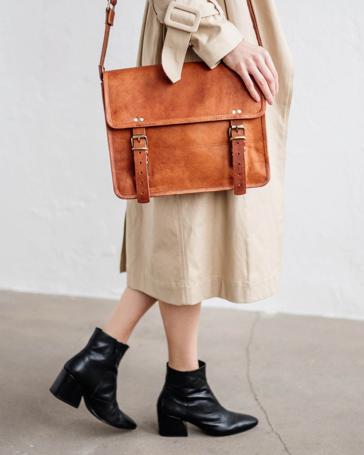 Cropped view of a woman wearing a beige trench coat and black leather heeled boots, carrying a vintage leather briefcase or satchel messenger bag with a shoulder strap against a white wall