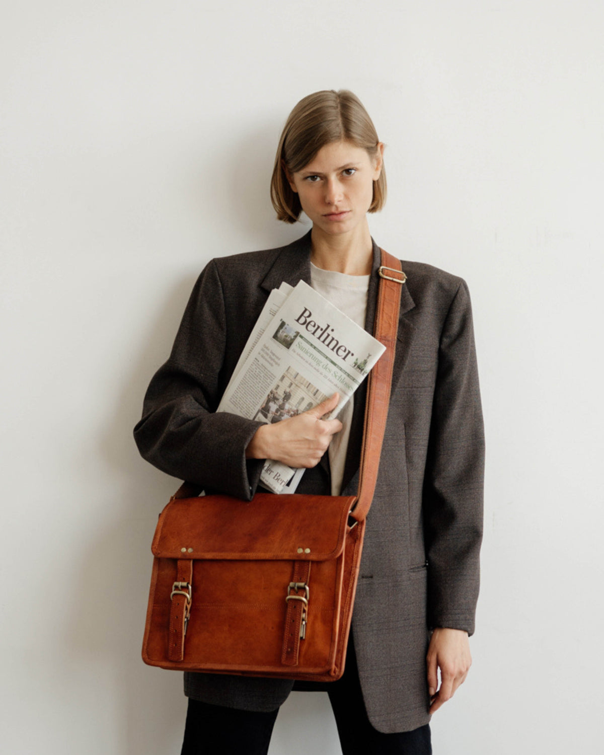 Three-quarter view of a woman with a bob haircut, wearing an oversized dark blazer, holding a folded German newspaper across her chest while carrying a vintage leather briefcase or satchel messenger bag via a shoulder strap against a white wall