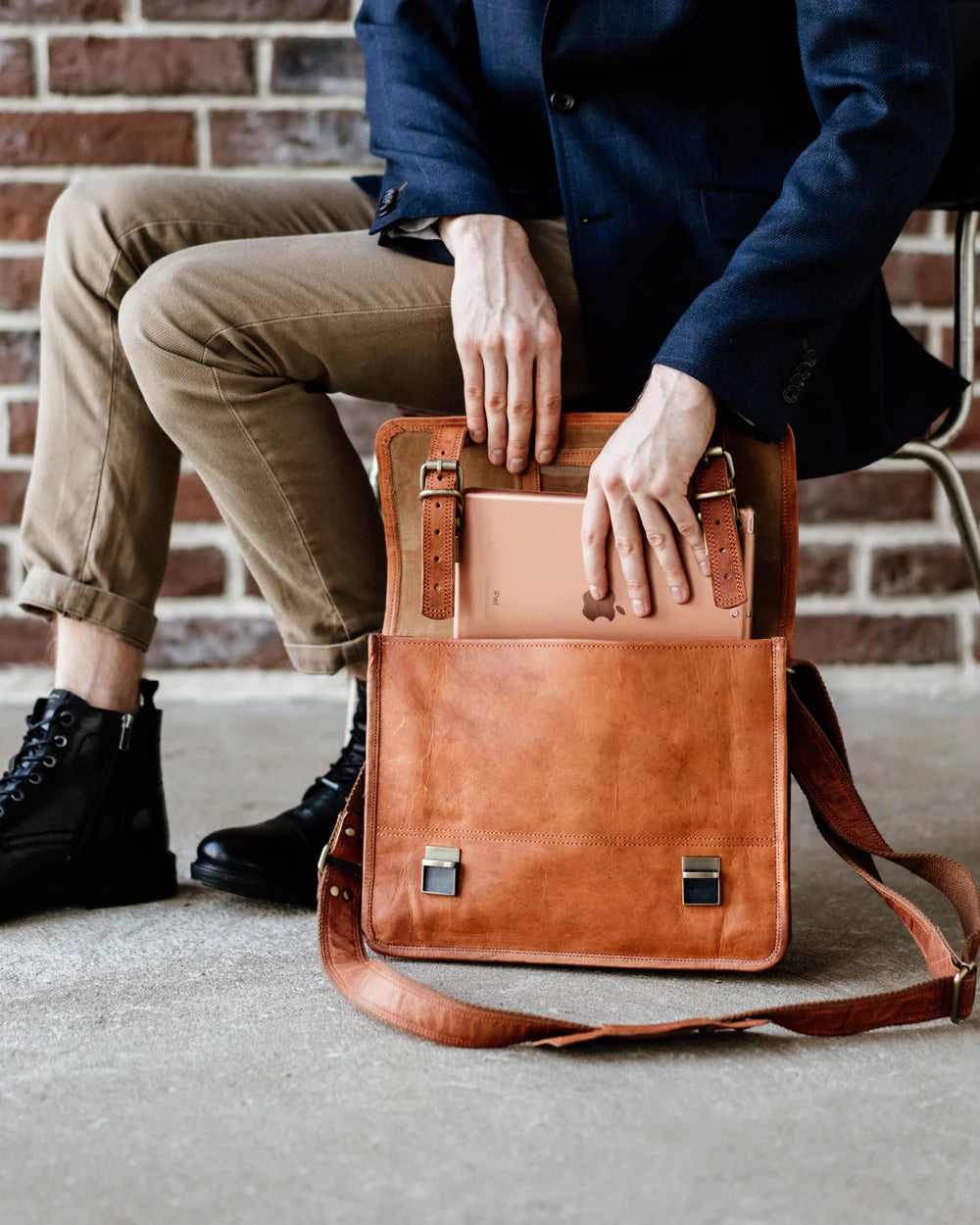 Cropped view of a seated man in a navy blazer and khaki pants, slide-inserting a tablet into an open vintage leather briefcase or satchel messenger bag resting on the ground