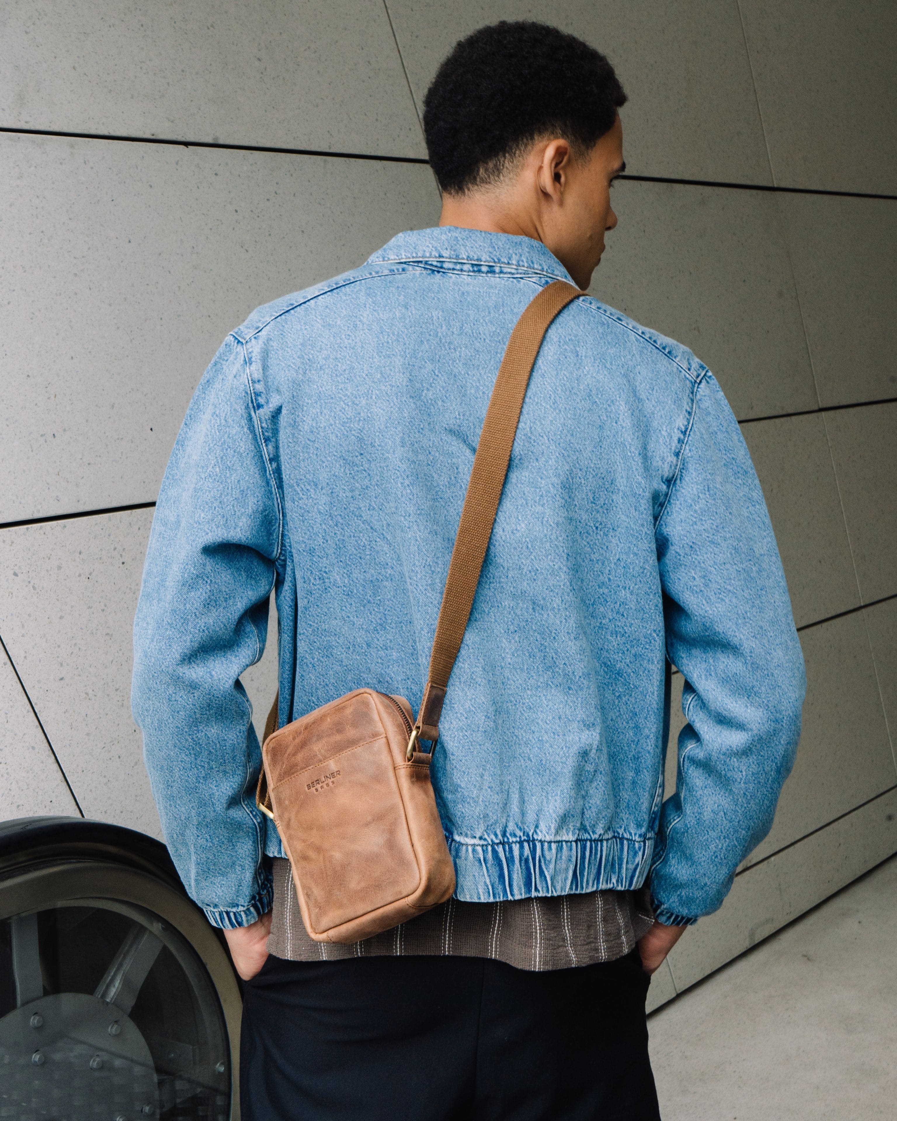 Rear view of man wearing a denim jacket and a camel leather messenger crossbody bag with a strap