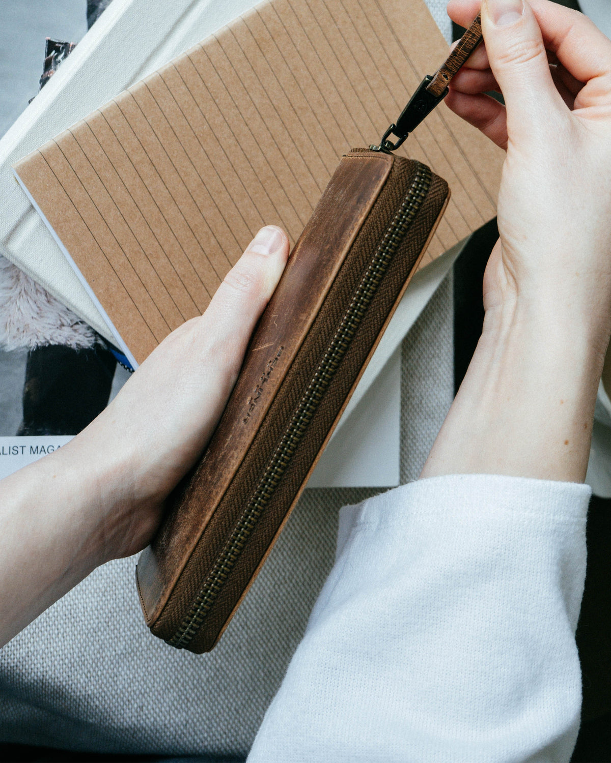 Person holding a walnut leather zip-around wallet, pulling on its leather-tabbed zipper.