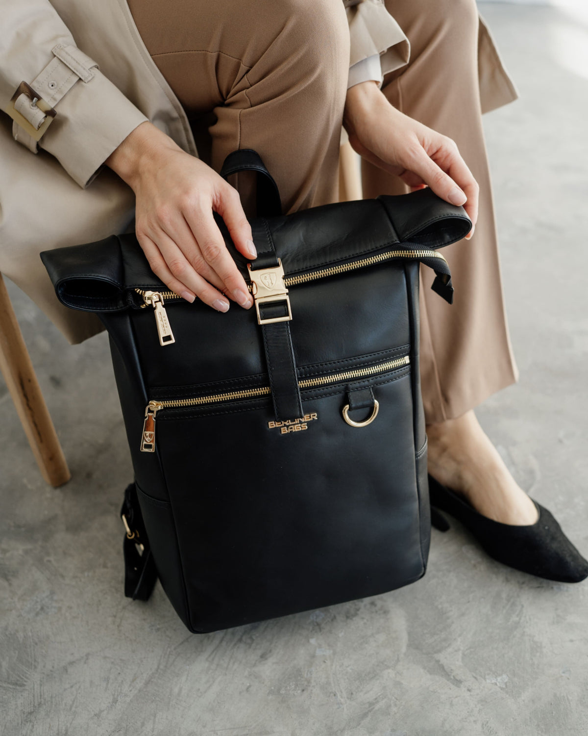 Person in trench coat holding a black roll-top backpack with gold hardware