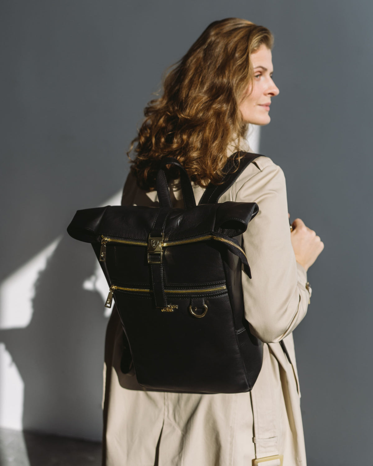 Woman in trench coat wearing black roll-top backpack with gold hardware