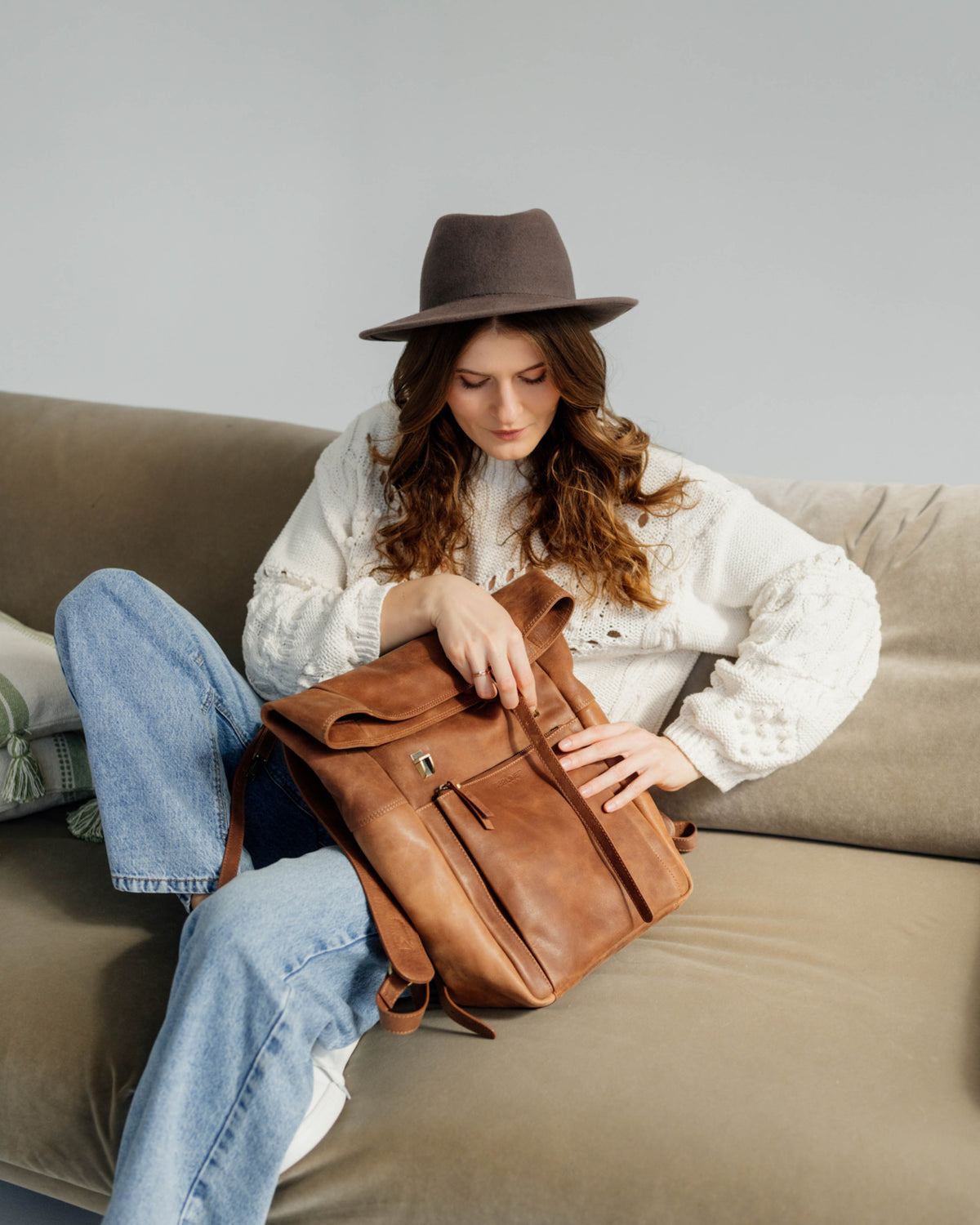 Lifestyle product shot of a woman on a sofa adjusting a camel roll-top backpack