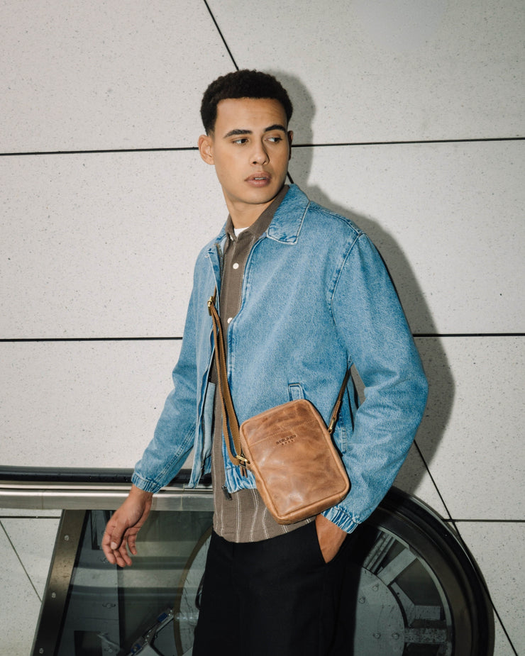 Angled front view of man looking away, wearing a denim jacket and a camel leather messenger crossbody bag with a strap
