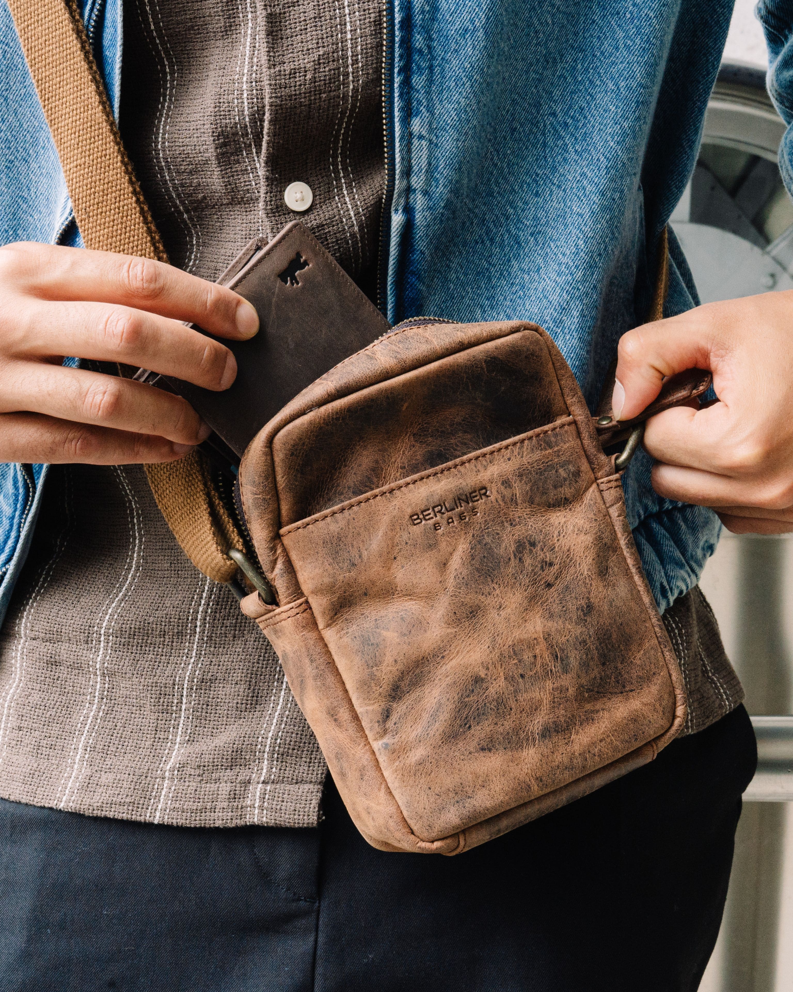 Close up view of man wearing a denim jacket and taking out a wallet from a earth leather messenger crossbody bag